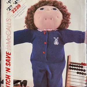 May include: A sewing pattern for a blue knitted outfit for a 16-inch doll. The pattern is called "Stitch 'n Save by McCall's" and the number is 9286. The doll is wearing the outfit and is holding a wooden abacus.