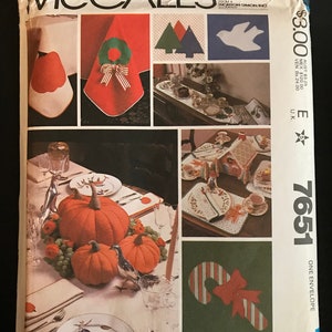 May include: A McCall's sewing pattern envelope with the number 7651. The pattern is for a variety of holiday-themed table decorations, including a pumpkin centerpiece, a candy cane, a Christmas tree, a dove, and a wreath. The envelope is white with red and green accents. The text "transfer included" is printed on the bottom left corner of the envelope. The text "PATTERN/PATRON" is printed on the bottom right corner of the envelope.