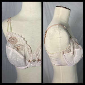 May include: A white lace bra with delicate floral embroidery and a pink trim. The bra has adjustable straps and a hook and eye closure in the back.