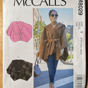 May include: McCall's M8029 sewing pattern featuring a hooded cape with a tie closure. The pattern includes illustrations of the cape in pink and black plaid. The image also shows a woman wearing a brown cape with a belt.