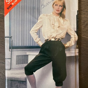 May include: A vintage sewing pattern for a blouse and pants. The pattern is for size B (14-16-18) and is from the See & Sew brand. The pattern features a woman wearing a white blouse with a ruffled collar and black pants.