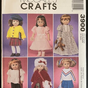 May include: McCall's Crafts sewing pattern for doll clothes. The pattern includes five different outfits: a yellow jacket with a plaid skirt, a pink dress, a floral dress, a red coat with a white hat, and a blue and white cheerleader outfit. The pattern number is 3900.