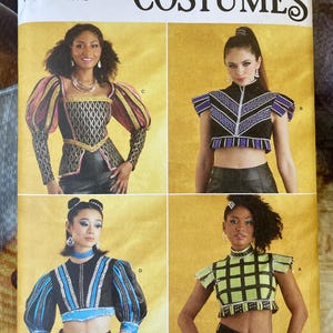 May include: Simplicity Costumes sewing pattern featuring four different costume designs. The pattern includes sizes U.S. 6-14. The costumes include a variety of tops with unique sleeve and neckline designs, in black, gold, pink, and blue.