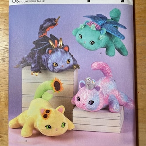 May include: Simplicity sewing pattern S9971 featuring four whimsical stuffed animal cat designs. The cats are in various colors, including yellow, blue, and pink, with decorative elements like wings, crowns, and sunflowers. The pattern is designed and printed in the USA.
