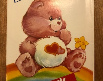 May include: A vintage sewing pattern featuring Love-a-Lot Bear from the Care Bears. The pattern is for a pink bear with a rainbow and a yellow flower. The pattern is by Butterick and is number 6232.