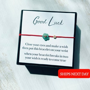 Puede incluir: Pulsera de cuerda roja con un dije de flor verde y un dije dorado. La pulsera está en una pequeña caja con el texto "Good Luck" e instrucciones sobre cómo hacer un deseo.