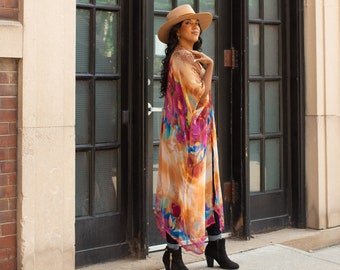 May include: A woman wearing a long, flowing floral kimono with a wide-brimmed hat. She is standing in front of a large, black, arched window.