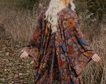 May include: A woman wearing a floral print kimono with a brown base color, accented with blue, orange, and yellow flowers. The kimono has long sleeves and a fringed hem. She is wearing blue jeans and tan boots, standing in a wooded area.