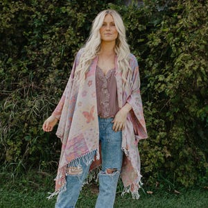 May include: A woman wearing a patterned shawl with butterfly and floral designs in pink, purple, and beige. She is also wearing a mauve top, ripped blue jeans, and brown ankle boots. The shawl has a fringed edge.