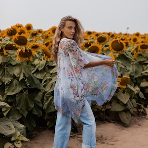 May include: A light blue floral print kimono with wide sleeves. The kimono is worn with light blue jeans. The background is a field of sunflowers under a cloudy sky. The kimono features a delicate floral pattern in shades of pink, purple, and green.