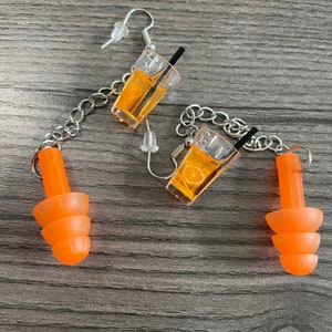 May include: A pair of orange silicone earplugs and a pair of earrings featuring a clear glass charm with a depiction of an orange drink with a straw and a slice of orange. The earrings are connected by a silver chain.