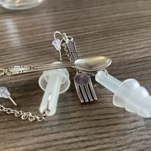 May include: A pair of silver earrings with a spoon, fork, and chain. The earrings are attached to clear plastic earplugs.