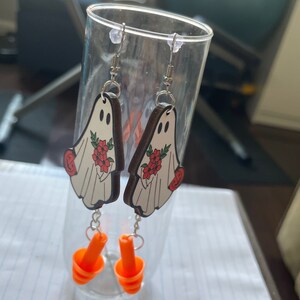 May include: A pair of wooden ghost earrings with red flowers and a white background. The earrings are hanging from silver hooks and have orange earplugs attached to the bottom.