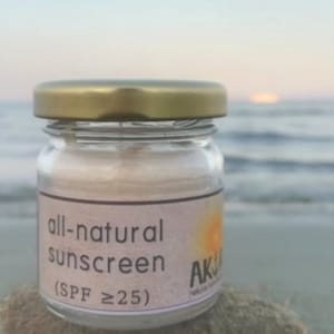 Op de afbeelding: Een kleine doorzichtige glazen pot met een goudkleurig metalen deksel met daarin een lichtgekleurde crème. Het etiket op de pot bevat de tekst all-natural sunscreen (SPF ≥25) en een zon-afbeelding. De pot staat op een zanderige ondergrond.
