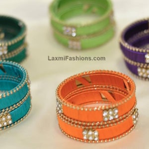 Può includere: Cinque set di braccialetti in filo di seta colorati con finiture dorate e accenti di strass. I braccialetti sono in tonalità di verde, arancione, viola e turchese.
