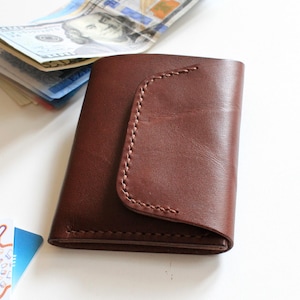 Może przedstawiać: Brązowy skórzany portfel z zakrzywioną klapką i przeszyciami. Portfel leży obok stosu banknotów i kilku kart kredytowych, wszystko na białej powierzchni.