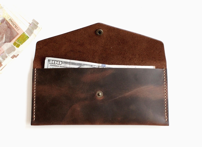 May include: A dark brown leather envelope wallet with a pointed flap, secured by a central metal snap button. The wallet features visible light-colored stitching along the sides and contains a one hundred dollar bill tucked inside the main compartment.