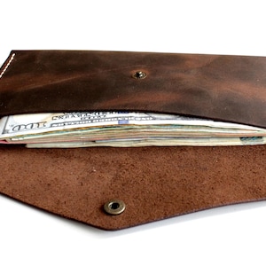 May include: A dark brown leather envelope-style wallet featuring a pointed flap secured with a single metal snap button. The wallet is partially open, revealing a stack of United States one hundred dollar bills tucked inside the main compartment.