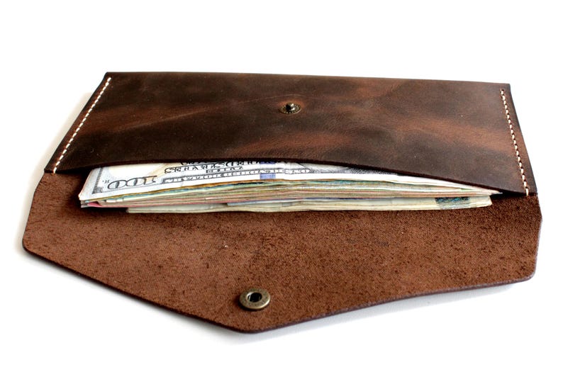 May include: A dark brown leather envelope-style wallet featuring a pointed flap secured with a single metal snap button. The wallet is partially open, revealing a stack of United States one hundred dollar bills tucked inside the main compartment.
