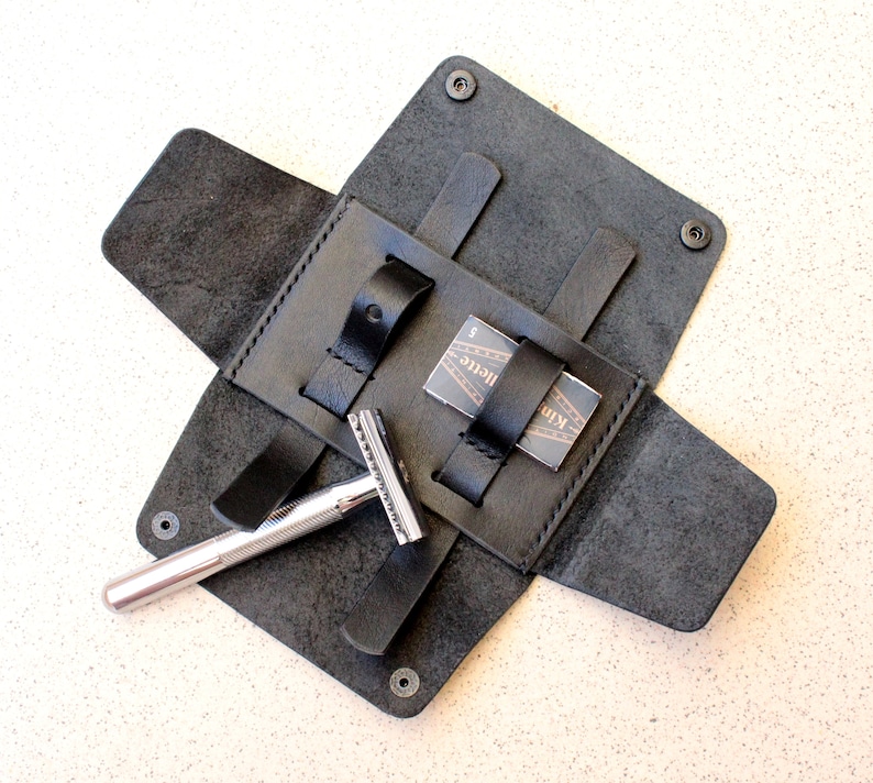 May include: A black leather travel case for a safety razor and razor blades. The case has a flap that folds over and closes with a snap. The case has a pocket for the razor and a separate pocket for the blades. The case is made of high-quality leather and is designed to be durable and long-lasting.