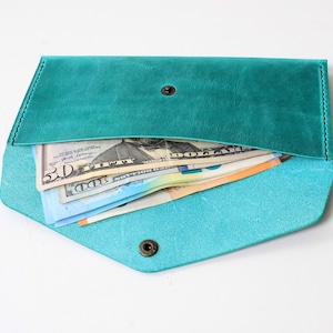 May include: A teal leather envelope wallet with a pointed flap and a single bronze-toned metal snap closure. The wallet is open and contains several paper banknotes, including a fifty-dollar bill and a one-hundred-dollar bill, visible inside.