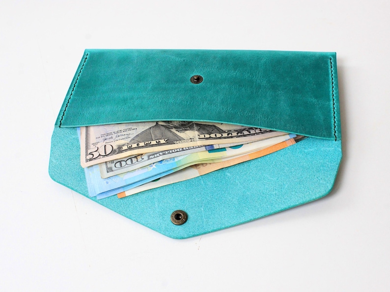 May include: A teal leather envelope wallet with a pointed flap and a single bronze-toned metal snap closure. The wallet is open and contains several paper banknotes, including a fifty-dollar bill and a one-hundred-dollar bill, visible inside.