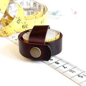 May include: A brown leather measuring tape holder with a brass snap button, holding a yellow measuring tape. A white and yellow measuring tape is partially unrolled, with a ruler in the foreground. Sewing pins are in the background.