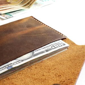 May include: A dark brown leather wallet with visible stitching along the top edge and a metal snap closure. The wallet is partially open, revealing a stack of United States one hundred dollar bills tucked inside the main compartment.