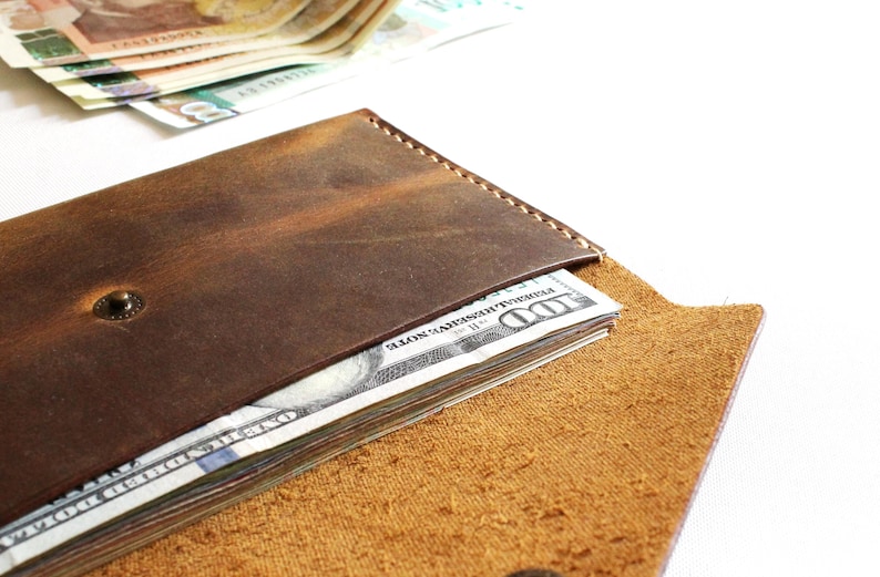 May include: A dark brown leather wallet with visible stitching along the top edge and a metal snap closure. The wallet is partially open, revealing a stack of United States one hundred dollar bills tucked inside the main compartment.