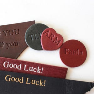May include: A collection of leather pieces featuring embossed text and shapes. Included are dark brown, black, and red leather strips and shapes with the phrases I LOVE YOU, ARM, Paola, and Good Luck printed in various fonts and styles.