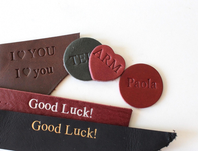 May include: A collection of leather pieces featuring embossed text and shapes. Included are dark brown, black, and red leather strips and shapes with the phrases I LOVE YOU, ARM, Paola, and Good Luck printed in various fonts and styles.