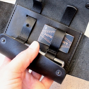 May include: A black leather case with a flap that opens to reveal a silver pen and a small card with the text "Kinfolk" and "Illete" printed on it.