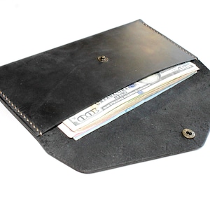 May include: A black leather envelope-style wallet featuring a pointed flap secured with a metallic snap button. The wallet is partially open, revealing a stack of United States one hundred dollar bills tucked inside the main compartment.