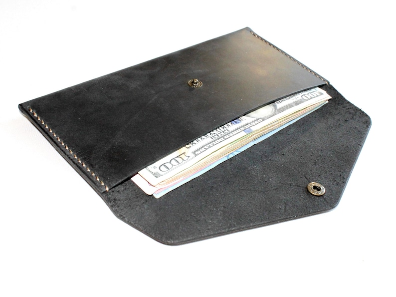 May include: A black leather envelope-style wallet featuring a pointed flap secured with a metallic snap button. The wallet is partially open, revealing a stack of United States one hundred dollar bills tucked inside the main compartment.
