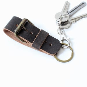 Puede incluir: Una correa de cuero marrón oscuro para llaves con una hebilla de metal color latón y una trabilla a juego. Un mosquetón de metal plateado está sujeto al extremo, sosteniendo un anillo de latón con tres llaves de metal plateado.