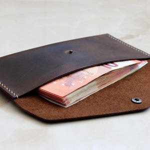 May include: A dark brown leather envelope wallet with visible light-colored stitching along the edges. The wallet features a single metal snap closure and contains a stack of orange ten-euro banknotes tucked inside the main compartment.