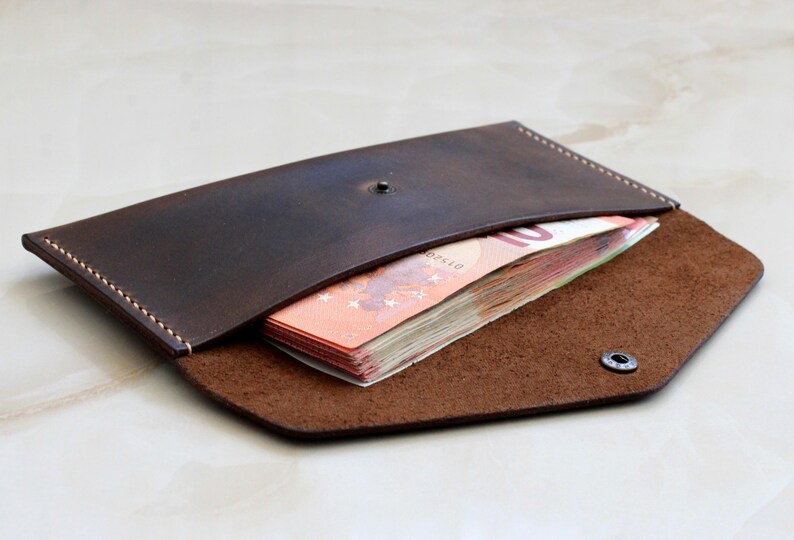 May include: A dark brown leather envelope wallet with visible light-colored stitching along the edges. The wallet features a single metal snap closure and contains a stack of orange ten-euro banknotes tucked inside the main compartment.