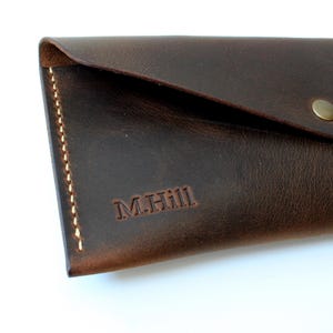 May include: A dark brown leather envelope-style wallet featuring a triangular flap secured with a single brass-colored metal snap button. The wallet is accented with visible light-colored stitching along the side and is embossed with M.Hill.