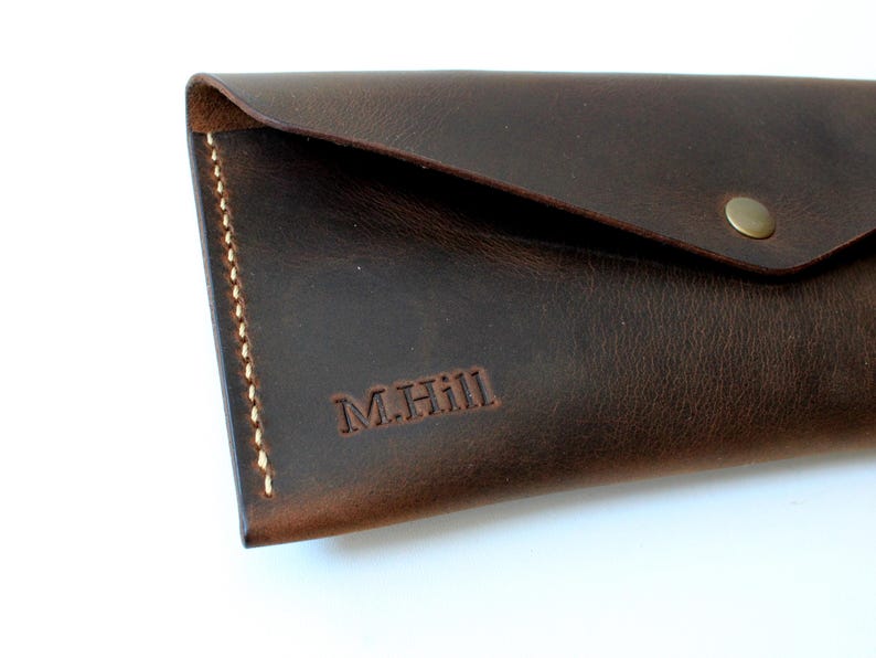 May include: A dark brown leather envelope-style wallet featuring a triangular flap secured with a single brass-colored metal snap button. The wallet is accented with visible light-colored stitching along the side and is embossed with M.Hill.