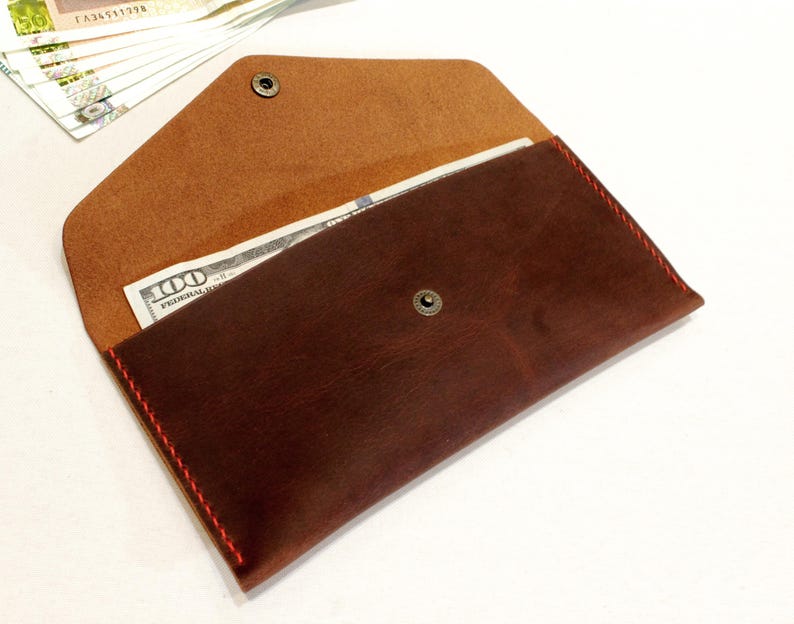 May include: A dark brown leather envelope wallet featuring a pointed flap with a metal snap closure. The wallet is stitched with vibrant red thread along the edges and contains a one hundred dollar bill inside the main compartment.
