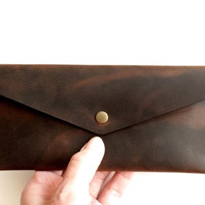 May include: A rectangular dark brown leather wallet with a pointed envelope-style flap closure. The wallet features visible light-colored stitching along the side edges and a single centered brass-toned metal snap button fastener on the front.