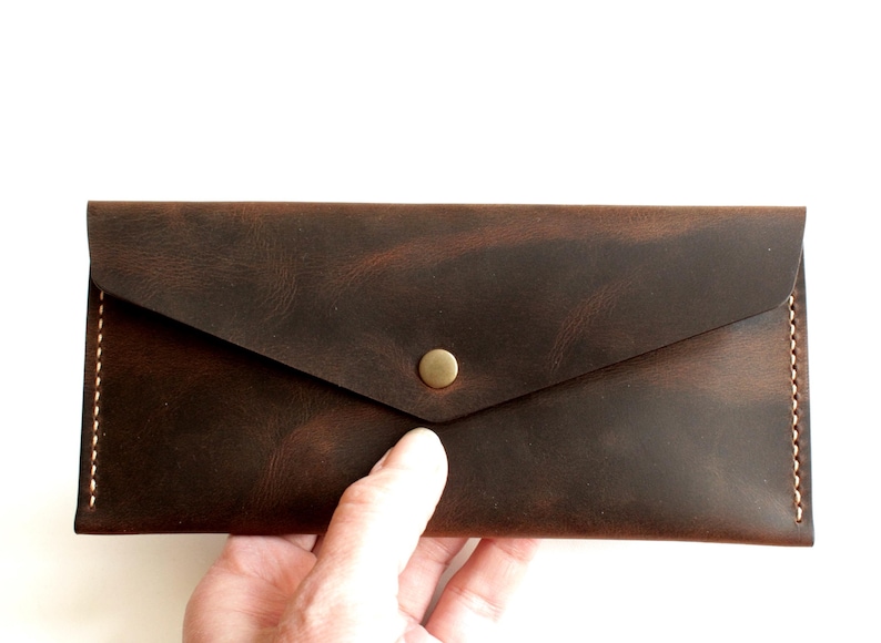 May include: A rectangular dark brown leather wallet with a pointed envelope-style flap closure. The wallet features visible light-colored stitching along the side edges and a single centered brass-toned metal snap button fastener on the front.