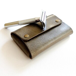 May include: A silver safety razor with a textured handle, resting on a brown leather travel case with brass snaps.