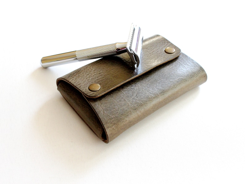May include: A silver safety razor with a textured handle, resting on a brown leather travel case with brass snaps.