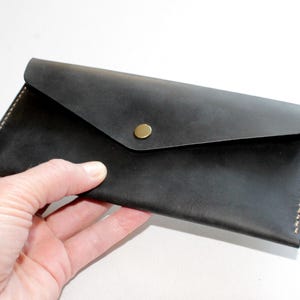 May include: A rectangular black leather envelope wallet featuring a pointed flap closure secured with a single brass-toned metal snap button. The wallet is constructed with visible light-colored stitching along the side edges and a smooth matte finish.