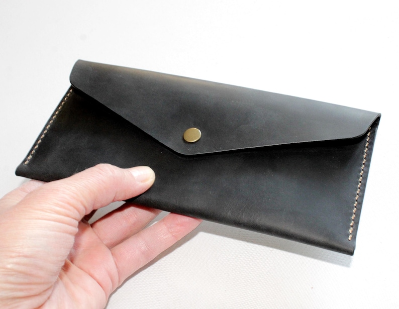 May include: A rectangular black leather envelope wallet featuring a pointed flap closure secured with a single brass-toned metal snap button. The wallet is constructed with visible light-colored stitching along the side edges and a smooth matte finish.