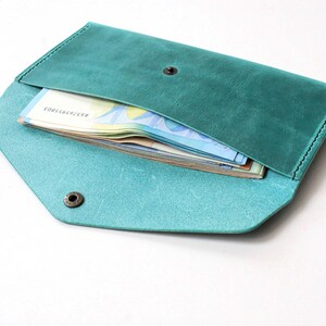 May include: A teal leather envelope wallet featuring a pointed flap, a dark metal snap button closure, and visible stitching along the edges. The wallet is partially open, revealing a stack of paper currency tucked inside the main compartment.