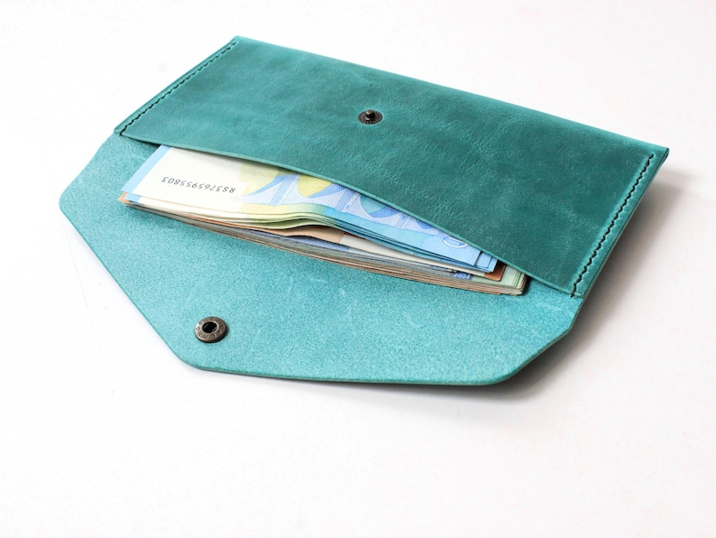 May include: A teal leather envelope wallet featuring a pointed flap, a dark metal snap button closure, and visible stitching along the edges. The wallet is partially open, revealing a stack of paper currency tucked inside the main compartment.