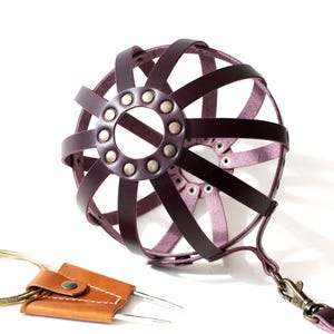 May include: A handcrafted, dark purple leather dog muzzle with a cage-like design. The muzzle features a circular front with rivets and a strap with a clasp. A small, tan leather case with metal needles is also present.