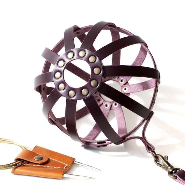 May include: A handcrafted, dark purple leather dog muzzle with a cage-like design. The muzzle features a circular front with rivets and a strap with a clasp. A small, tan leather case with metal needles is also present.